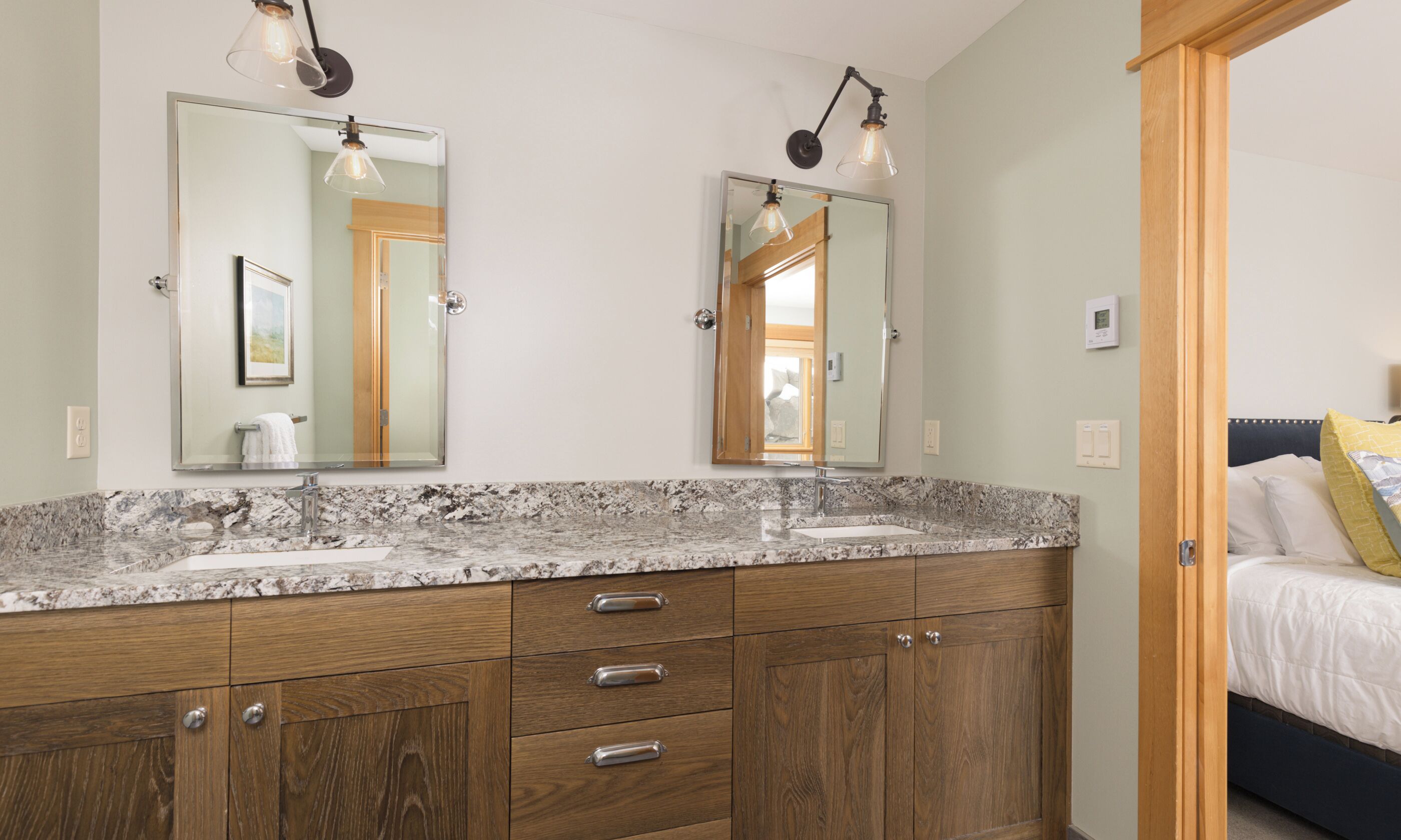 A bathroom with two mirrors, a granite countertop, wooden cabinets, and a bed visible through an open door.