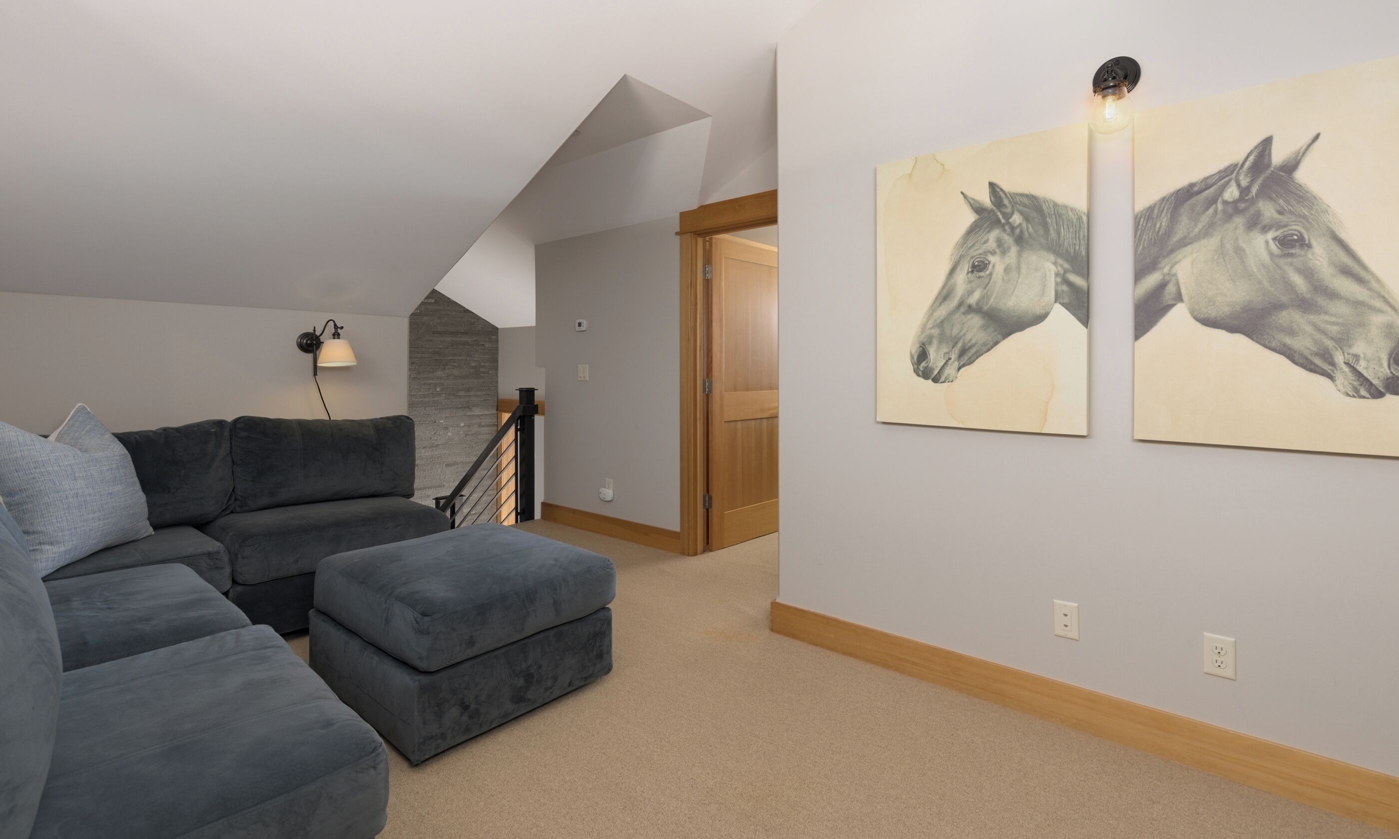 Un salon moderne avec un canapé d’angle gris, un petit pouf, un escalier et deux œuvres d’art murales sur le thème du cheval.