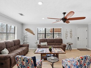 A modern living room with a brown sofa set, a wooden coffee table, patterned chairs, a ceiling fan, and large windows. - Lake Martin Adorable Lakefront Beauty (Dadeville)