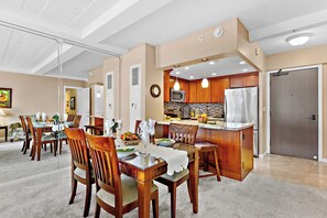 A modern kitchen with wooden cabinets, a stainless steel refrigerator, and a dining area with wooden chairs and a glass table. - Postcard Ocean View of Kaanapali ~ 1BD/1BA (Lahaina)