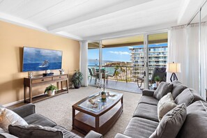 En stue med sofa, sofabord, fjernsyn og en balkon med udsigt over havet og bygninger.