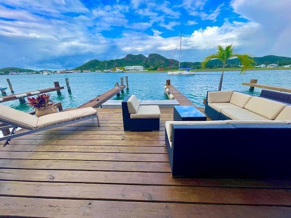Una terraza de madera con sillones de descanso y una pequeña mesa, con vista a una masa de agua con barcos y una costa lejana.