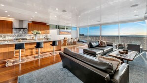 A modern kitchen with a marble countertop, wooden cabinets, and a large island with bar stools. There is a dining area with a table and chairs, and a living room with a leather sofa and a large window overlooking a scenic view.