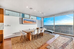 A modern dining area with a wooden table, white chairs, and a large window offering a view of the cityscape.