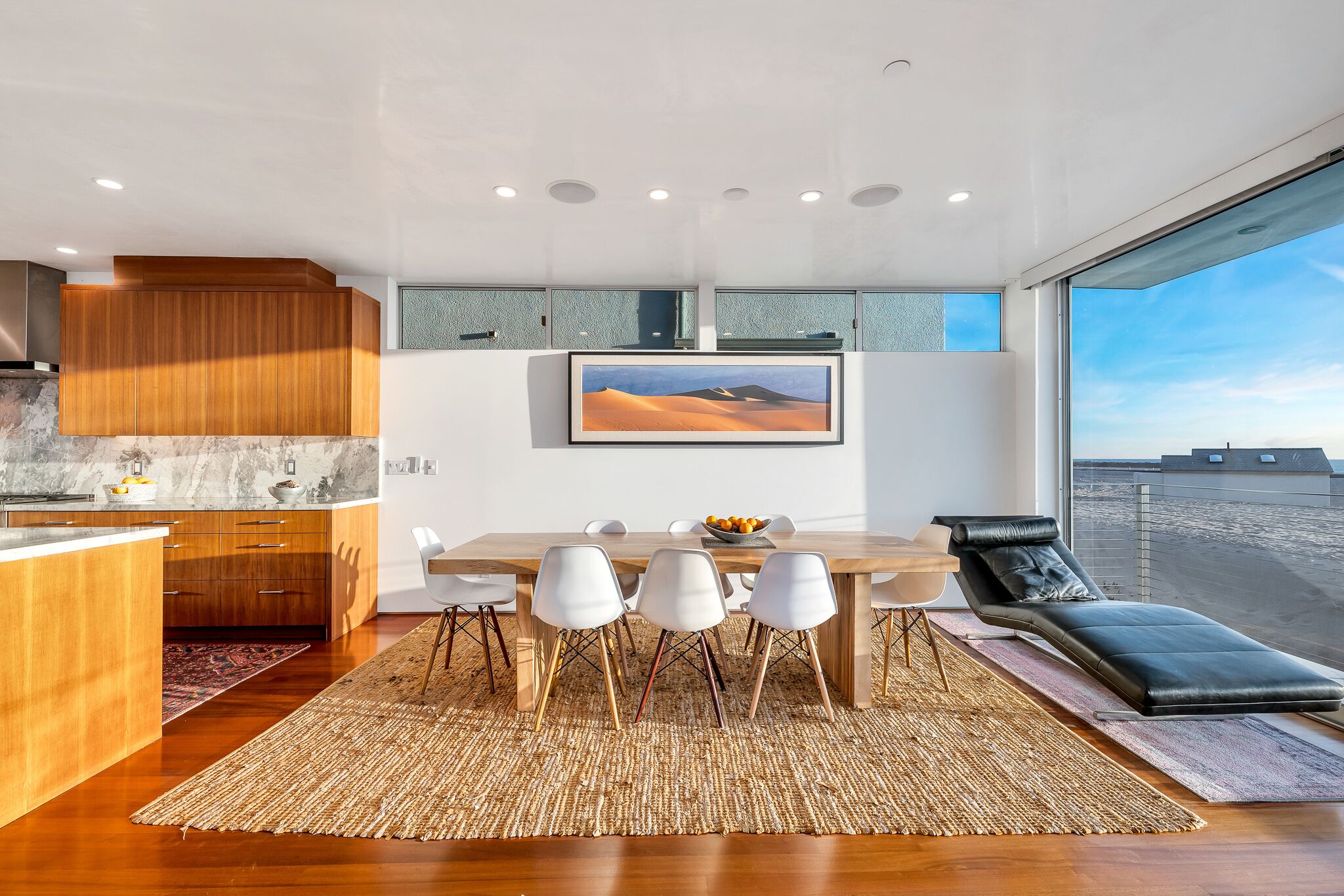 Una cocina moderna con armarios de madera, encimera de mármol y comedor con mesa de madera y sillas blancas.