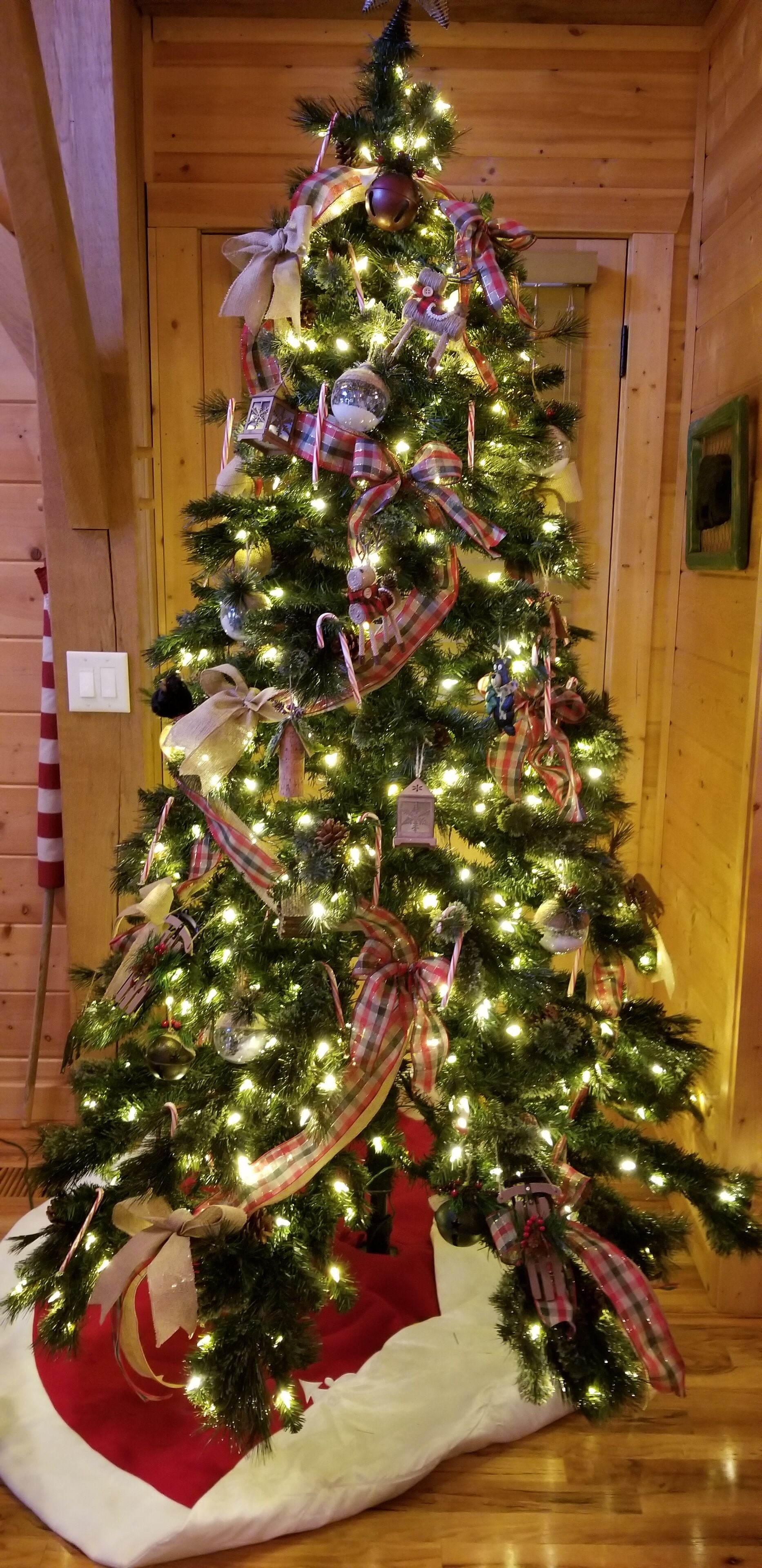 Ein mit Lichtern, Bändern und Schmuck verzierter Weihnachtsbaum in einem Holzraum mit Tür und einem gerahmten Bild an der Wand.