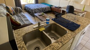 Une chambre d’hôtel compacte comprenant un coin cuisine, un lit avec une literie à motifs, un bureau avec une chaise et un téléviseur fixé au mur.