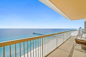 A balcony with a view of the ocean and a pier. There are white chairs and a table on the balcony.