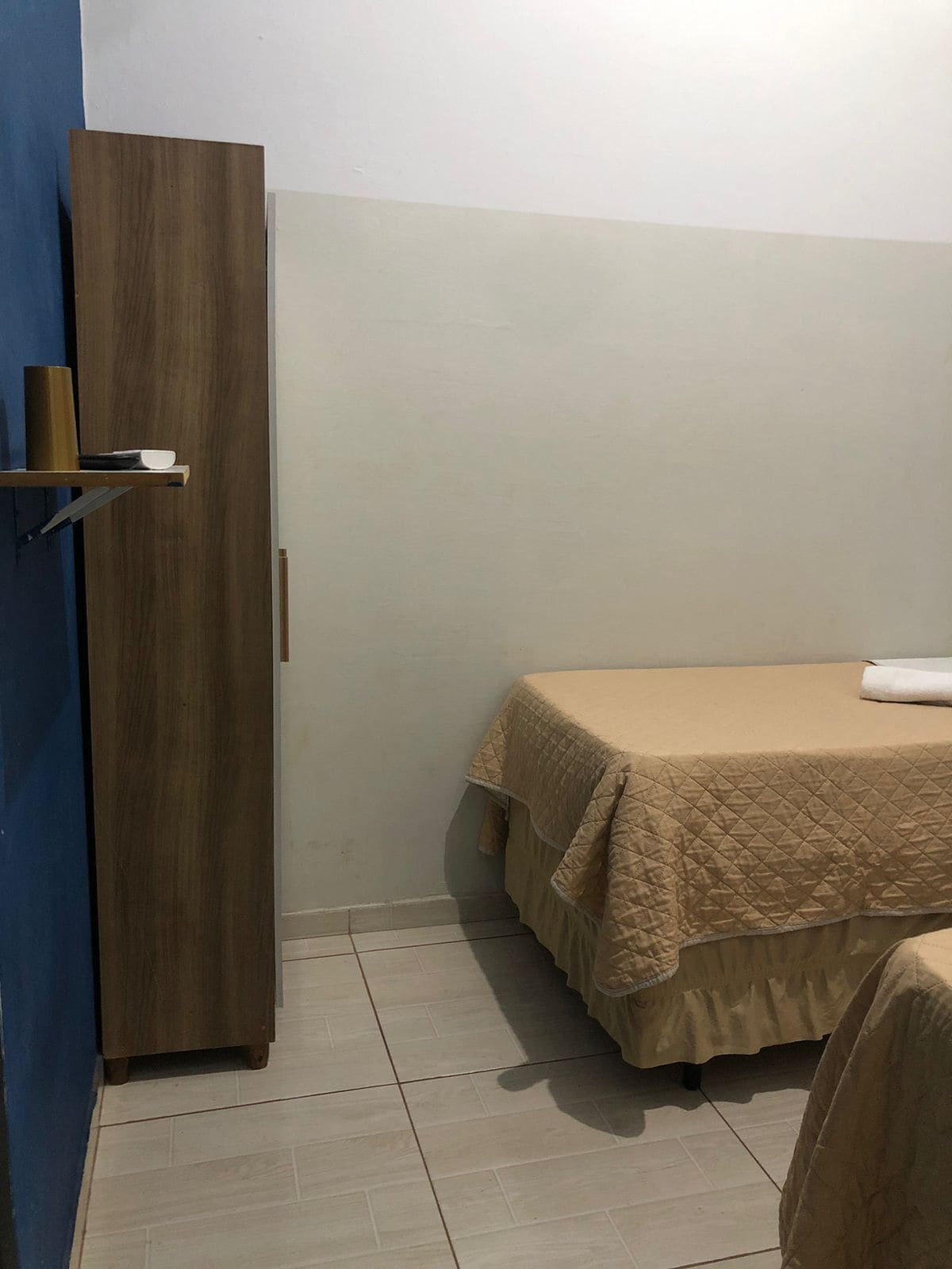 A small, neatly arranged room with a bed, a wooden wardrobe, and a shelf with a roll of paper towels.