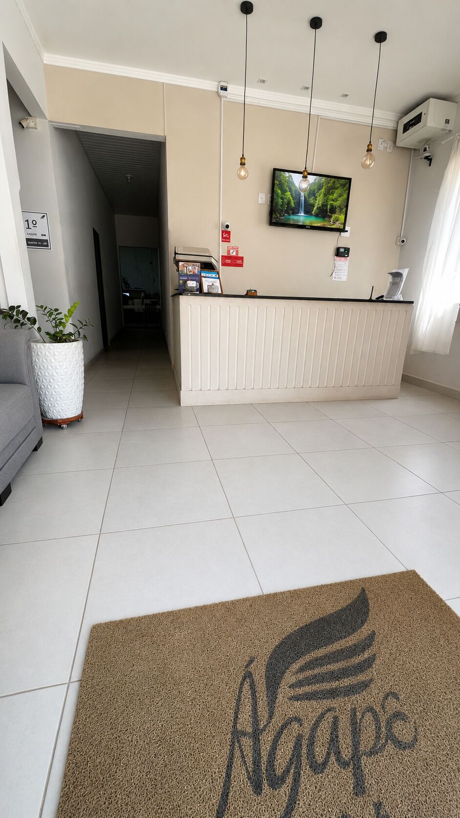 Zona de recepción de un hotel, con mostrador de recepción, televisión montada en la pared y felpudo con el nombre del hotel.