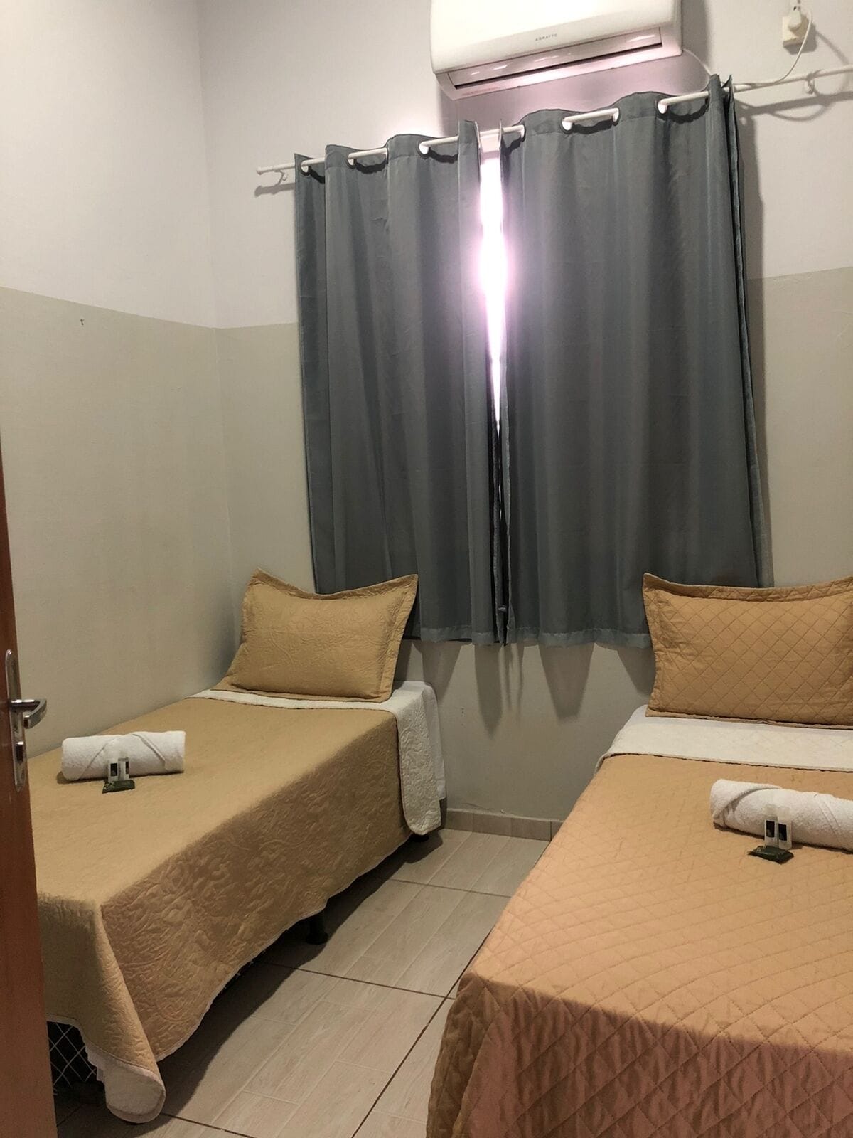 Habitació d'hotel amb dos llits individuals, cadascú amb un llençol beix i una almoçada. Hi ha una finestra amb cortines grises i una unitat de aire condicionat muntada sobre la finestra.