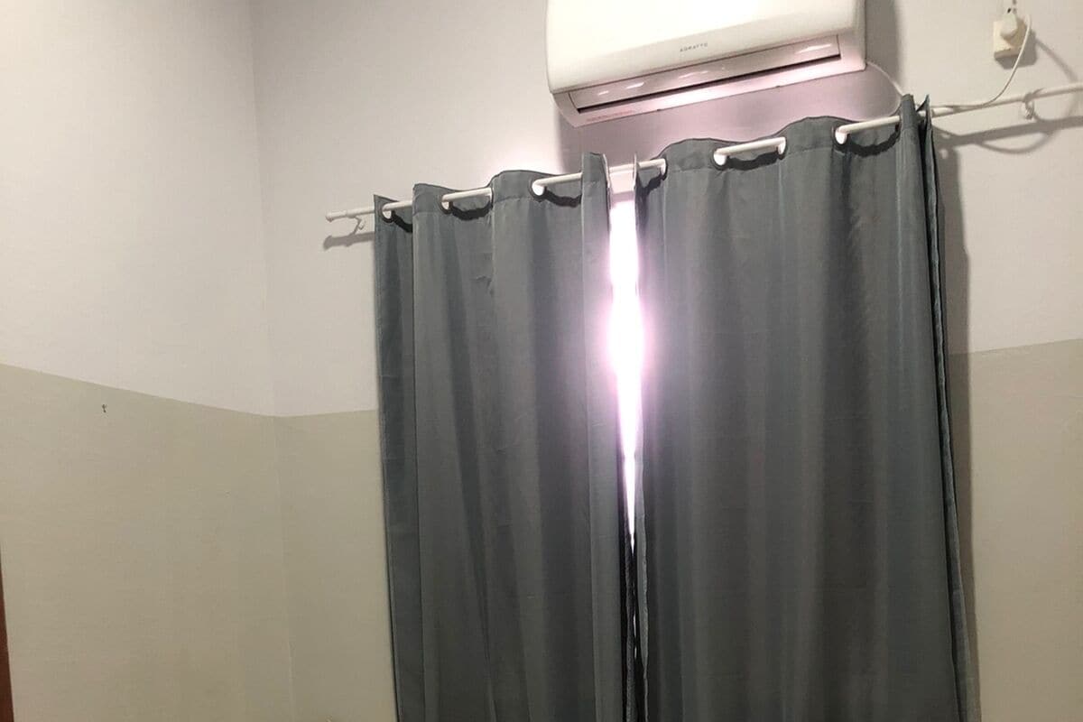 Quarto de hotel com duas camas de solteiro, cada uma com colcha bege e uma almofada. Há uma janela com cortinas cinzas, e um aparelho de ar condicionado montado acima da janela.