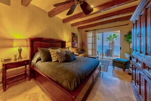 A bedroom with a large bed, a ceiling fan, a nightstand, a lamp, a painting, a window with a view of palm trees, and a sliding door leading to a balcony. - Villa LA Estancia LOS Cabos (CABO SAN LUCAS)