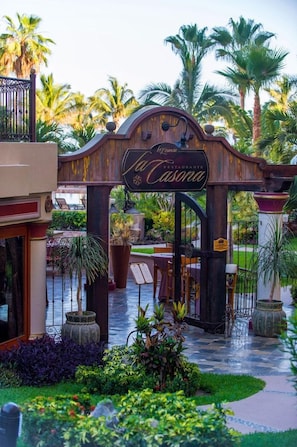 A restaurant entrance with a wrought iron gate, palm trees, and a well-maintained garden. - Villa LA Estancia LOS Cabos (CABO SAN LUCAS)