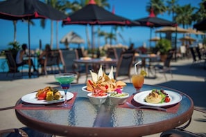 A dining table with various dishes and drinks, surrounded by outdoor seating and umbrellas, with a pool and palm trees in the background.
