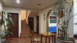 Una escalera que sube a un segundo piso, una mesa de madera con una silla, y una planta grande en maceta.