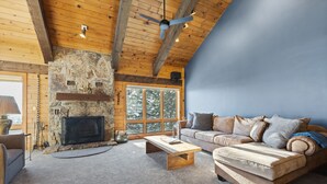 A cozy living room with a stone fireplace, a wooden ceiling, and a large window offering a view of snow-covered trees. - Less Than 100 Yards to Slopes + Private Hot Tub w/ Gondola Views (Steamboat Springs)