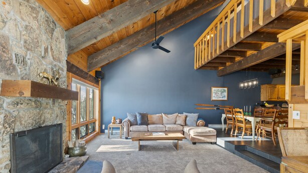 A spacious living room with a stone fireplace, a large sofa set, a dining area with wooden chairs, and a high ceiling with exposed beams. - Less Than 100 Yards to Slopes + Private Hot Tub w/ Gondola Views (Steamboat Springs)