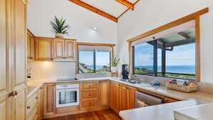 A modern kitchen with wooden cabinets, a built-in oven, and a large window offering a view of the sea. - Zenith - Freycinet Holiday Houses (Coles Bay)