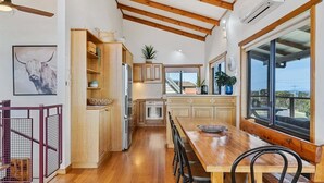 A modern kitchen with wooden beams, a dining area with a large window, and a view of the outdoors. - Zenith - Freycinet Holiday Houses (Coles Bay)