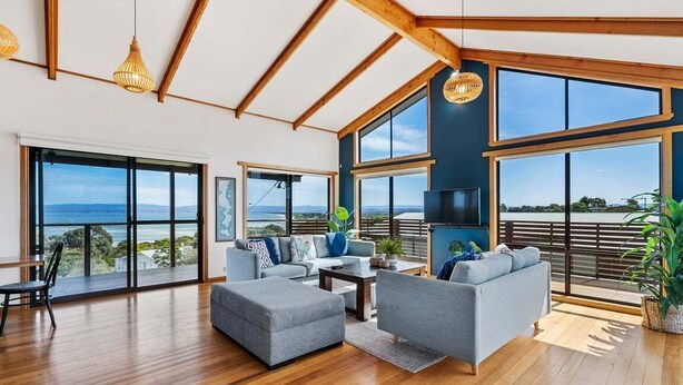 A spacious living room with a large window overlooking the sea, wooden flooring, and modern furniture. - Zenith - Freycinet Holiday Houses (Coles Bay)