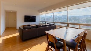 A modern living room with a sofa, a TV mounted on the wall, a dining table with chairs, and large windows offering a city view. - Capitalia | Near to ABC Hospital with Gym/Parking (Ciudad de México)