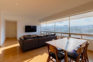 A modern living room with a large window, a flat-screen TV, a sofa, and a dining table with chairs.