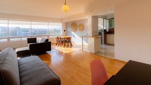 A modern living room with a sofa, dining table, and chairs, a kitchen area, and large windows offering a city view. - Capitalia | Near to ABC Hospital with Gym/Parking (Ciudad de México)