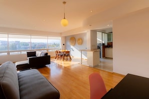 A modern living room with a sofa, dining table, and chairs, a kitchen in the background, and large windows offering a city view.