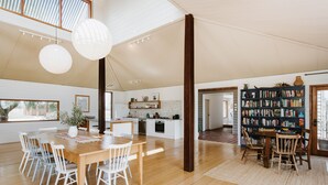 A spacious living area with a wooden dining table, white chairs, a bookshelf, and a kitchen in the background. - Abercorn - one of Stayz’s 2025 Holiday Homes of the Year (Buckaroo)