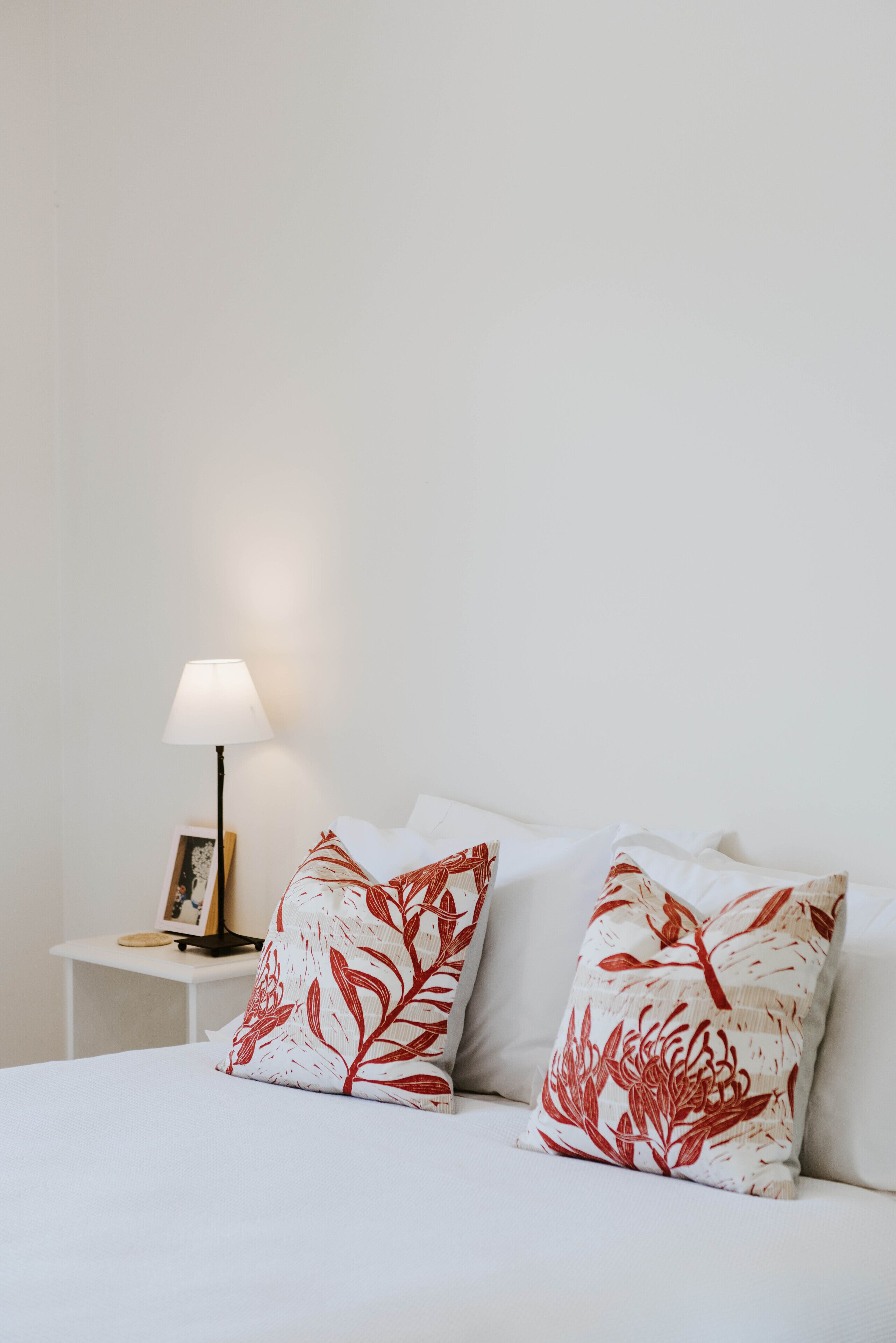 A neatly made bed with white bedding and two decorative pillows with red floral patterns. A bedside table with a lamp and a framed picture is visible.