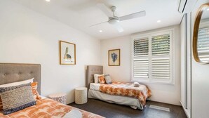 Ein Schlafzimmer mit zwei Betten, einem Deckenventilator, einem Fenster mit Jalousien und gerahmten Kunstwerken an der Wand.