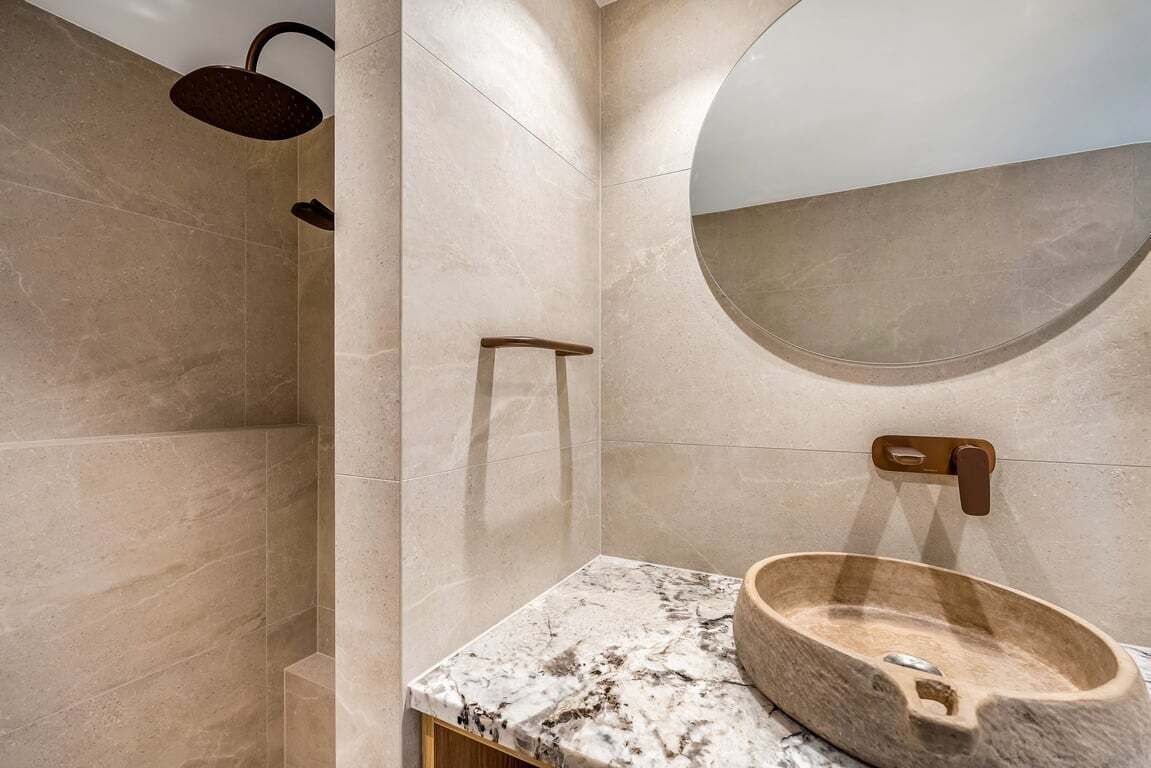 A modern bathroom with a marble countertop, a wooden basin, a wall-mounted mirror, and a shower with a circular overhead light.