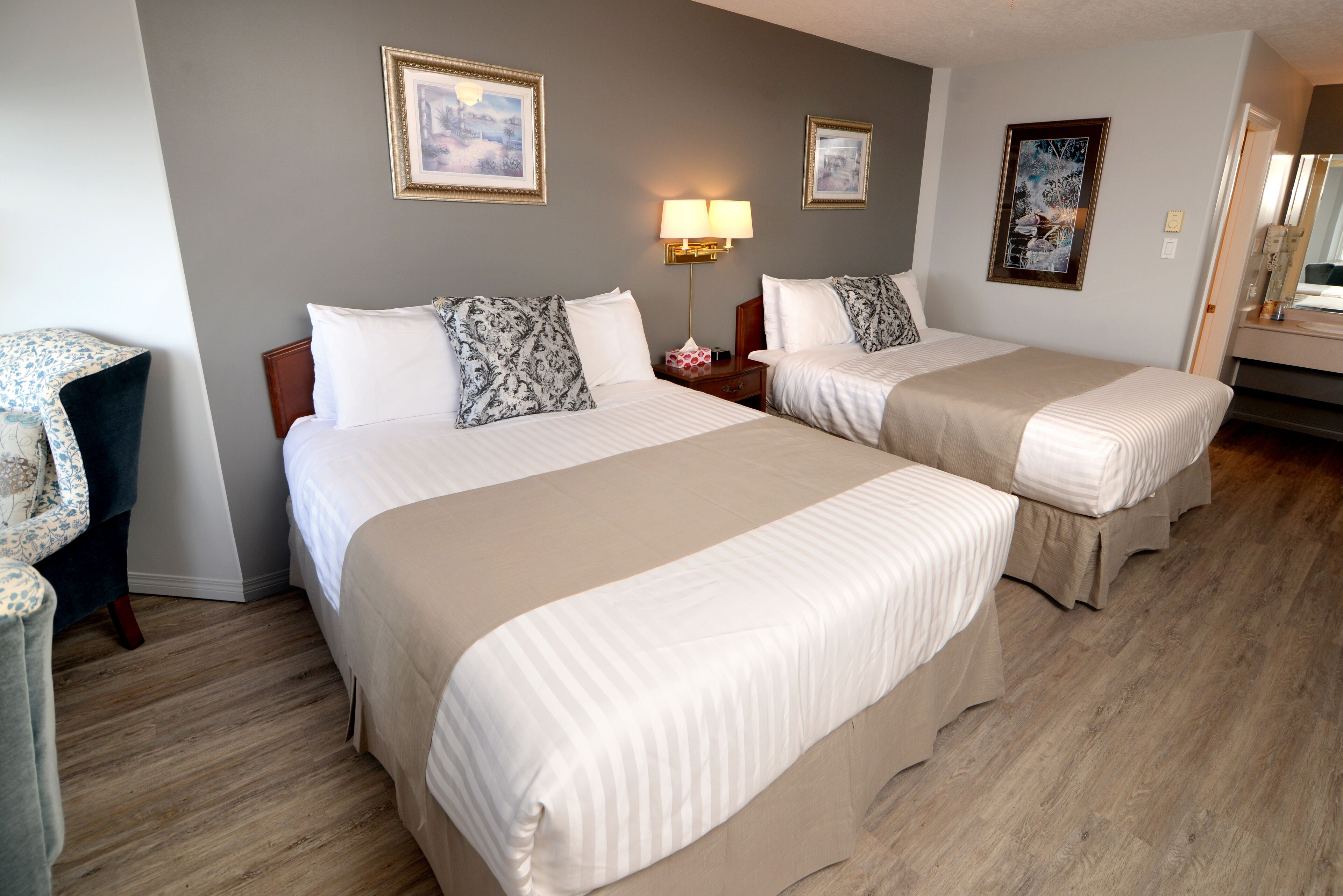 A hotel room with two beds, a nightstand with a lamp, and a chair.