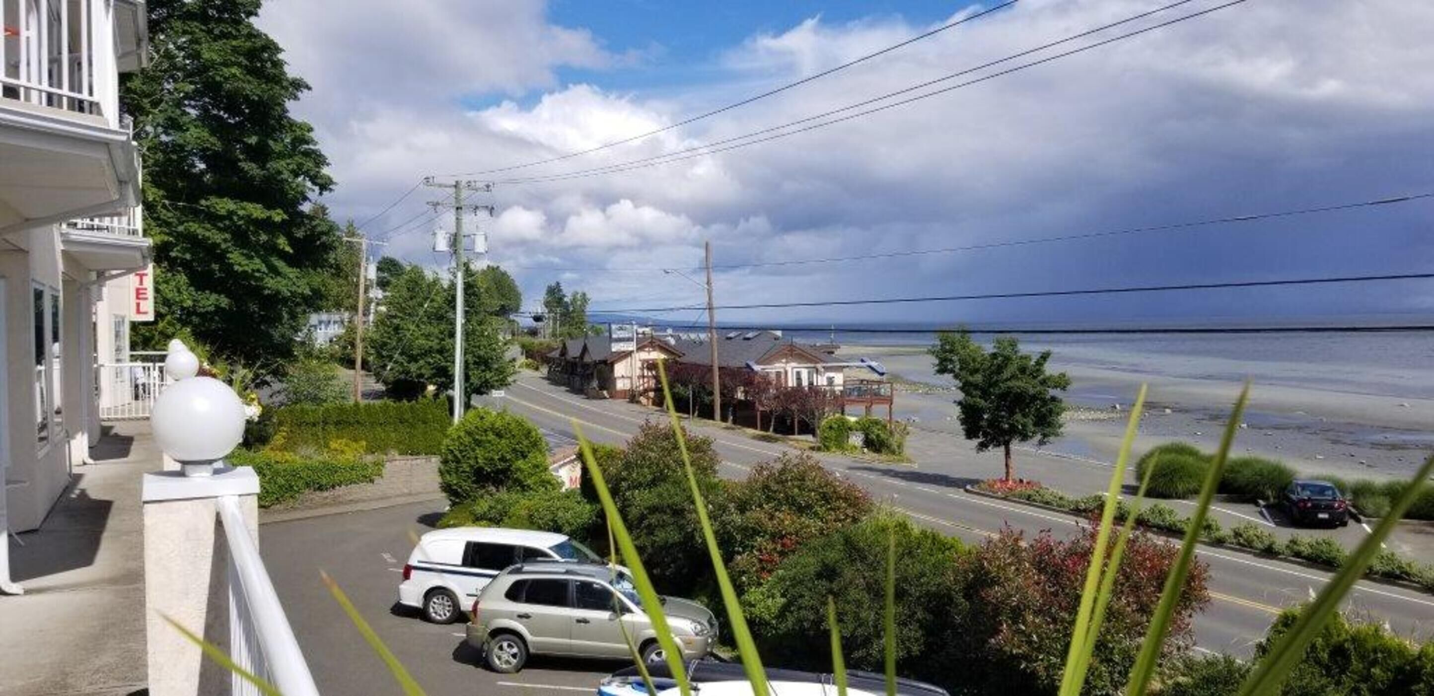 A coastal street with parked cars, a building with a balcony, and a view of the sea.