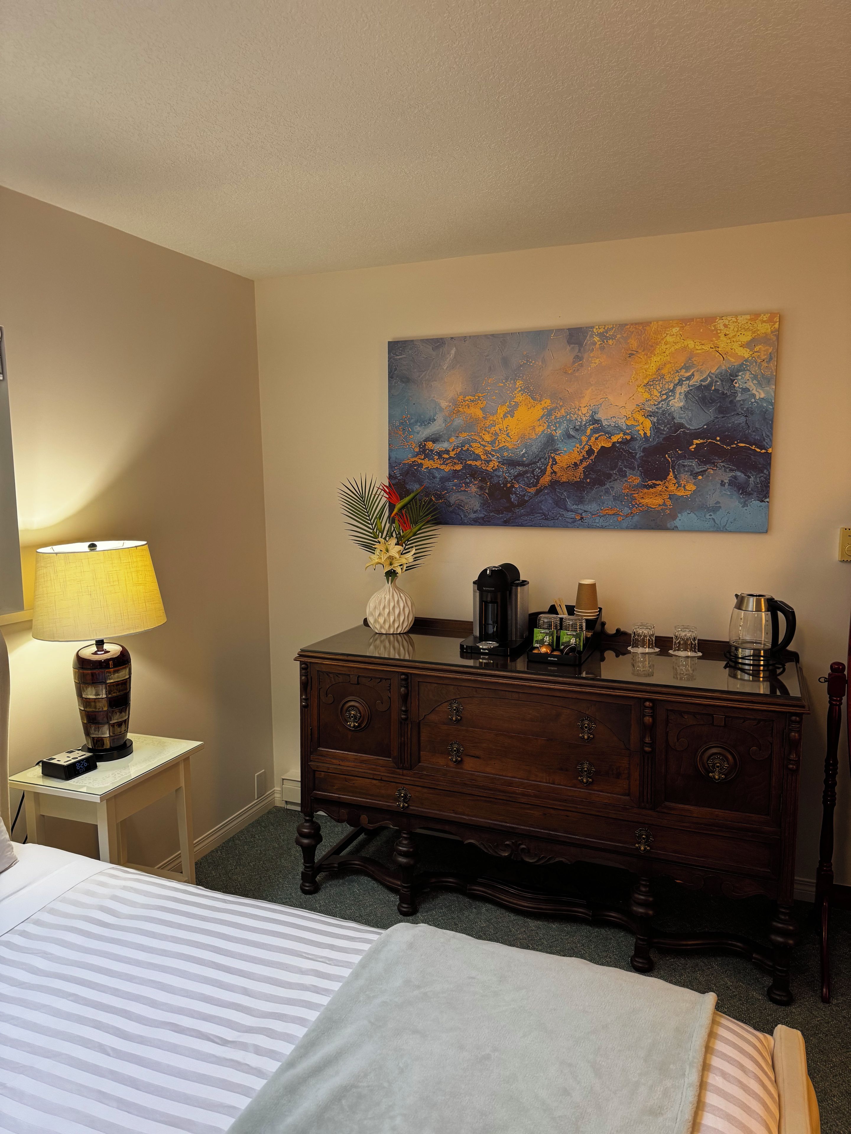 Habitación de hotel con cama, mesita de noche con lámpara, cómoda con varios objetos y un cuadro en la pared.