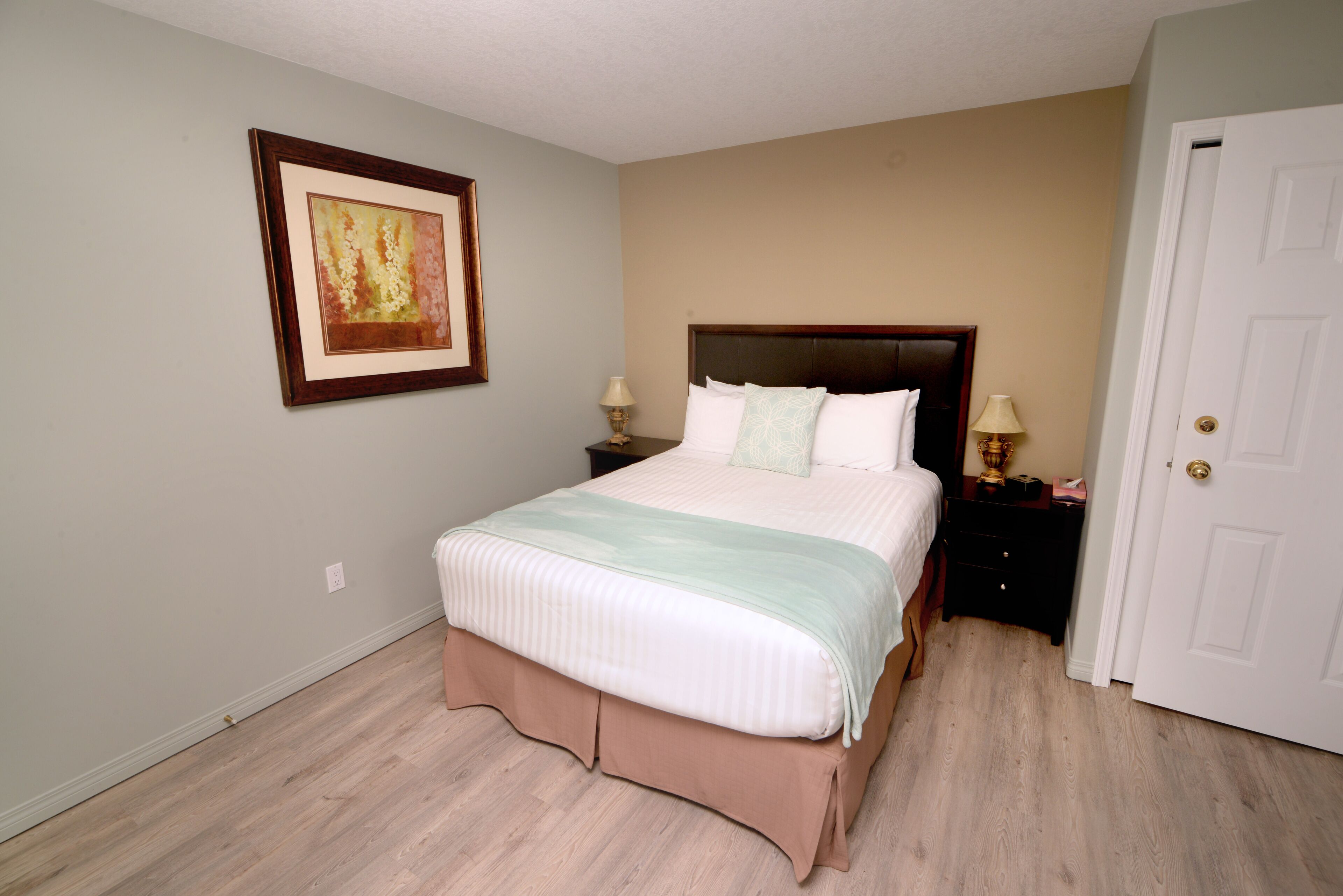 Un dormitorio con cama, dos mesitas de noche, un velador, una puerta y un cuadro enmarcado en la pared.
