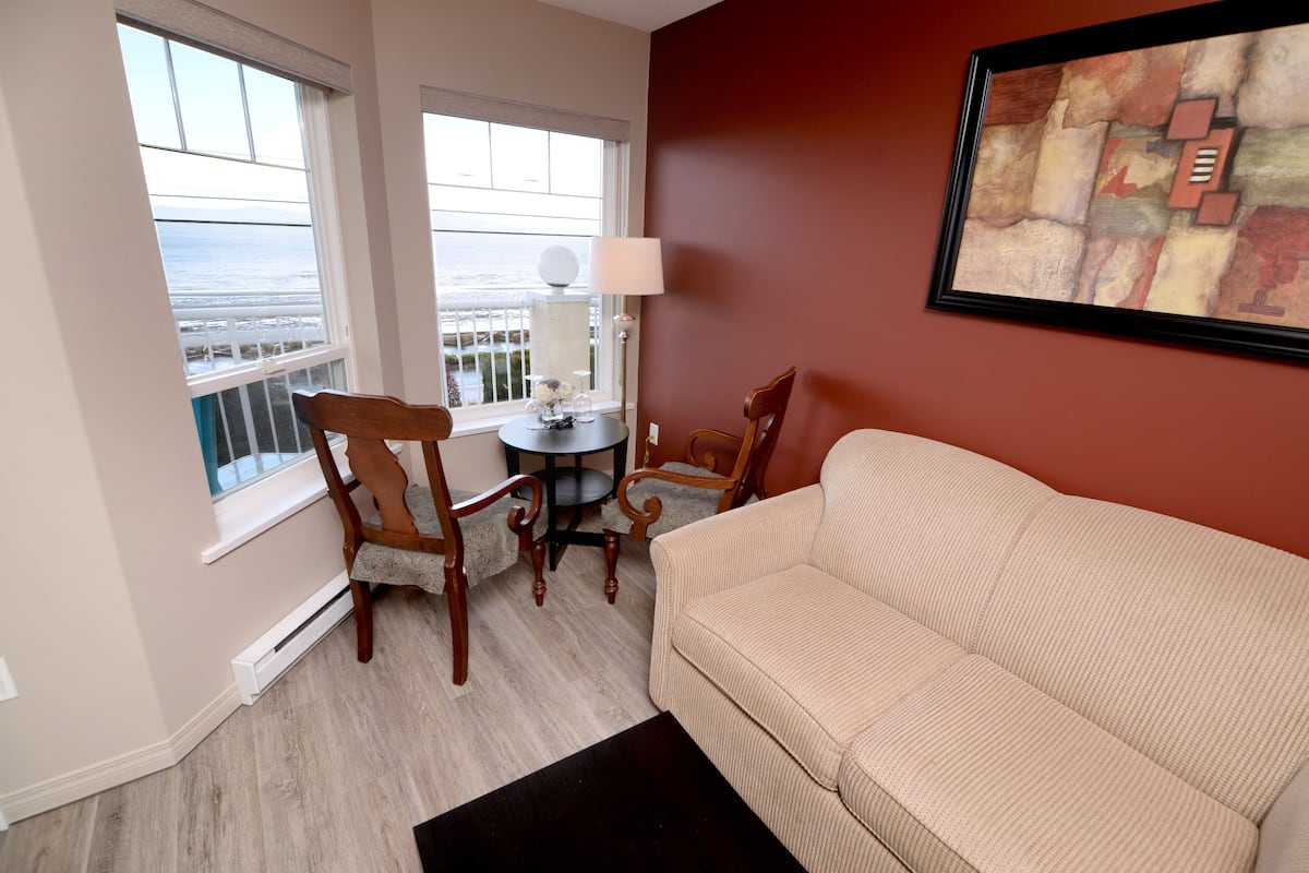 A room with a beige sofa, a wooden chair, a small round table with a lamp, and a view of the ocean through large windows.