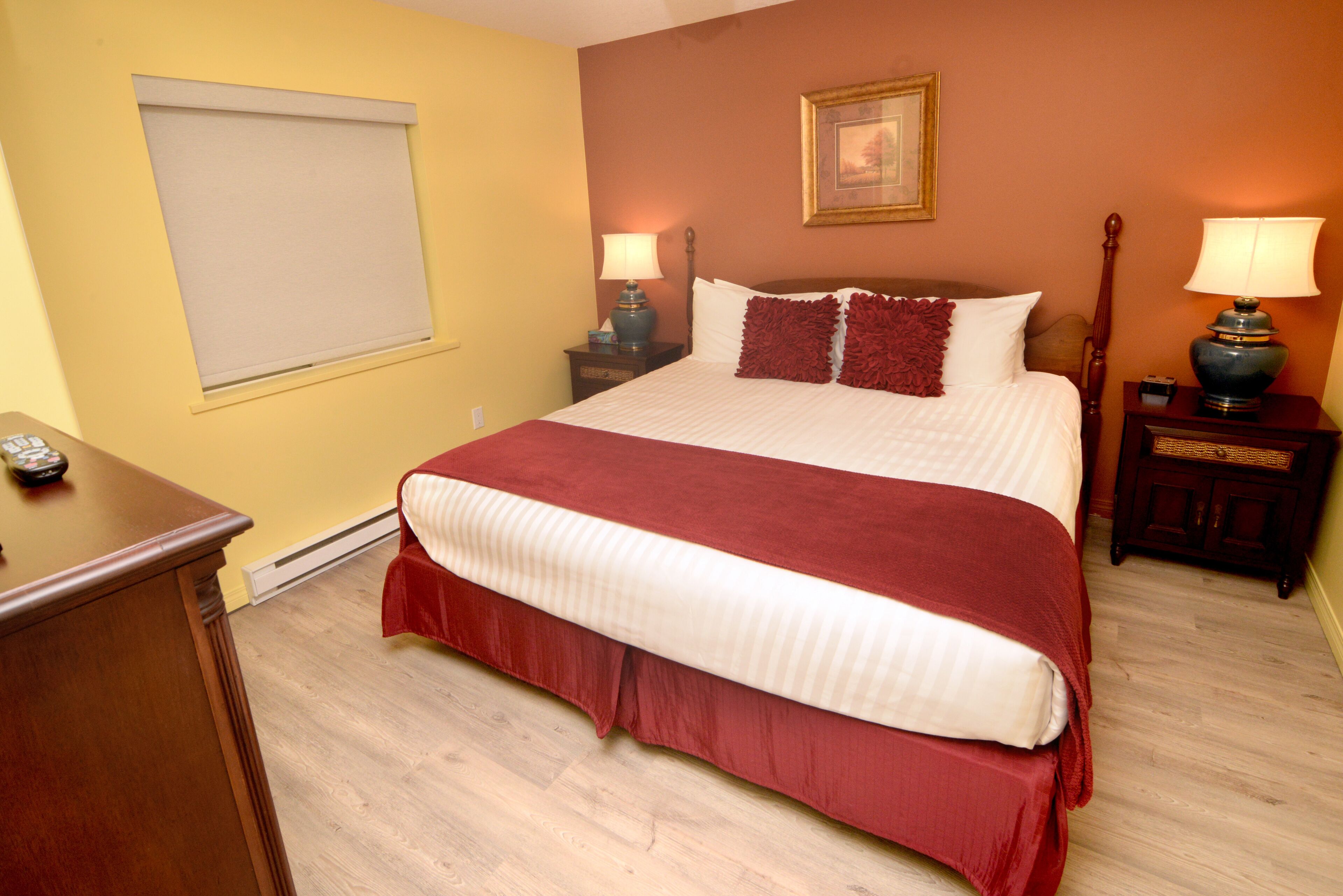 A bedroom with a large bed, two bedside tables with lamps, a wooden dresser, a window with a blind, and a framed picture on the wall.