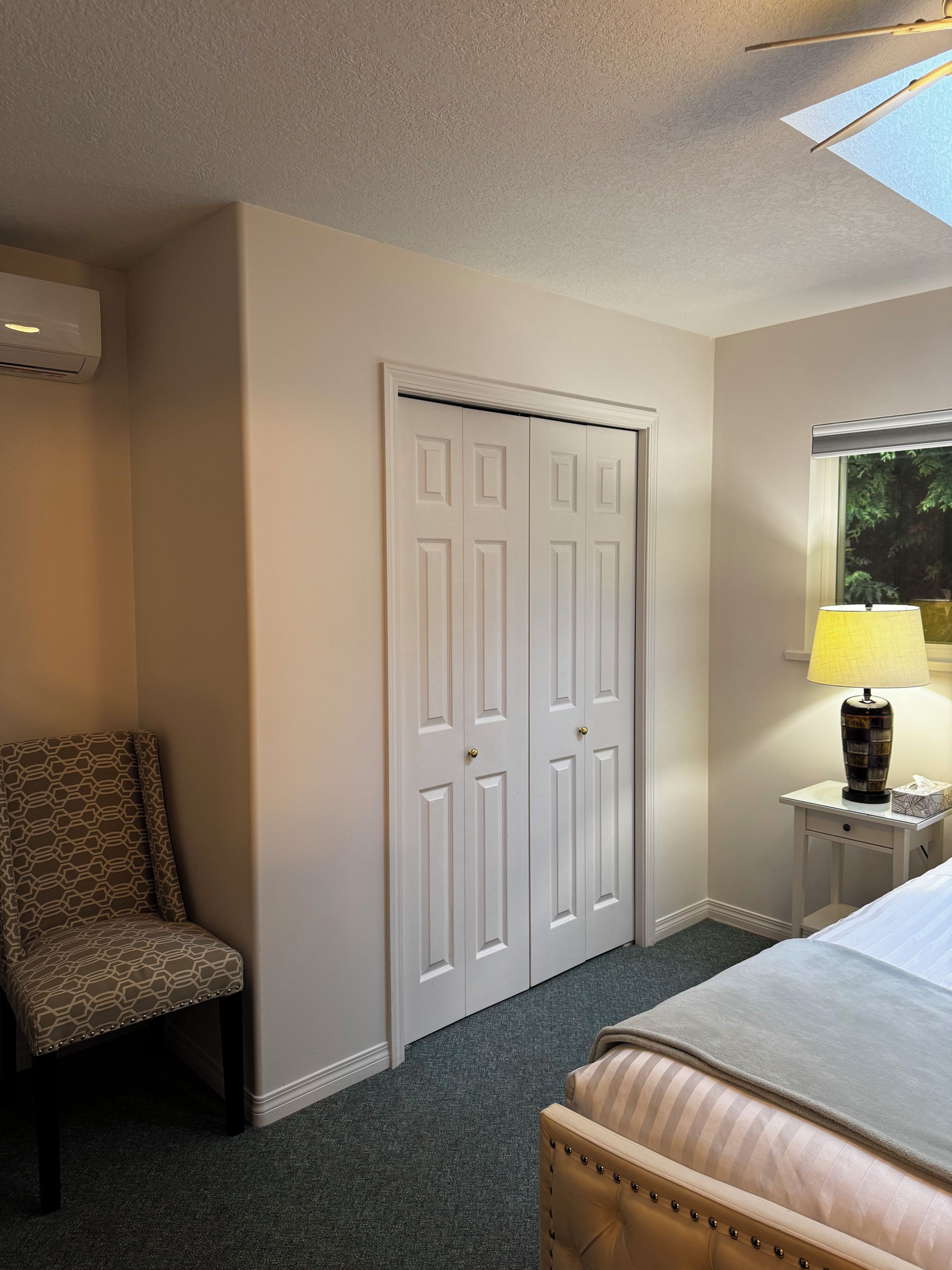 A hotel room with a bed, a chair, a desk with a lamp, and a wardrobe.