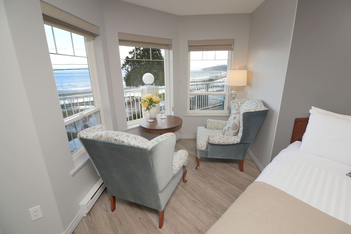A hotel room with a bed, two armchairs, a small table with a vase of flowers, and a balcony with a railing and a view of the sea.