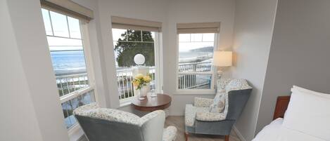 Habitación de hotel con cama, dos sillones, una mesita con un florero y balcón con barandilla y vista al mar.