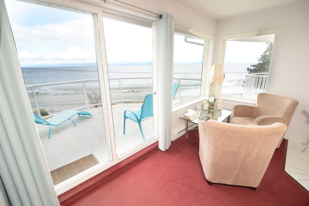 A room with a large window overlooking a beach, a red carpet, a beige armchair, and a glass table with a vase.