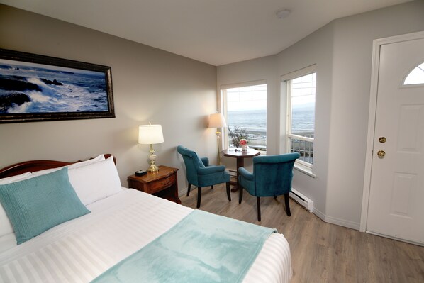 A hotel room with a bed, two chairs, a table, a lamp, a painting, and a view of the ocean through large windows.