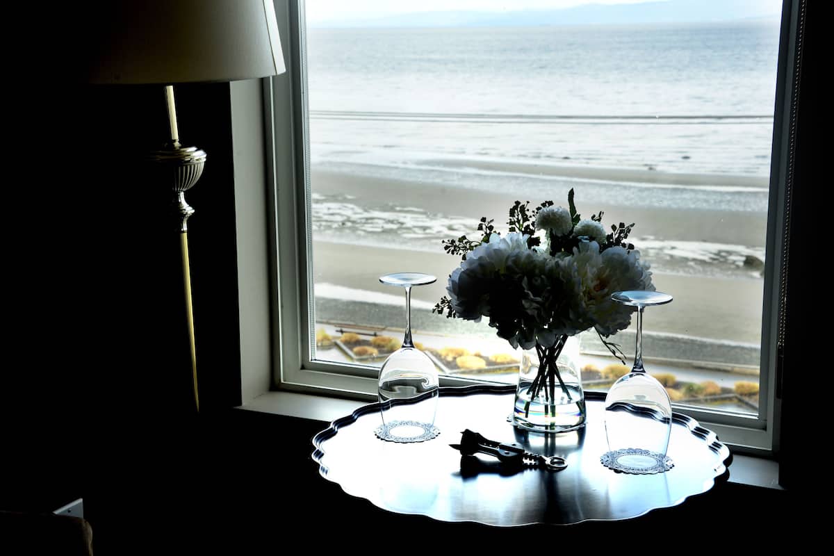 A table with a vase of flowers, a wine glass, and a bottle opener by a window with a view of the beach.
