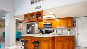 A kitchen with wooden cabinets, a stainless steel refrigerator, a microwave, and a granite countertop. There are two bar stools and a potted plant on the counter. - The Octagon at Capricorn (South Padre Island)
