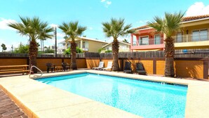 A swimming pool with lounge chairs and palm trees, surrounded by a wooden fence and buildings in the background. - The Octagon at Capricorn (South Padre Island)