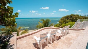 Eine Terrasse mit weißen Stühlen und einem Tisch, mit Blick auf das klare, blaue Meer, verbunden durch einen gewundenen Steinweg und umgeben von Grün.