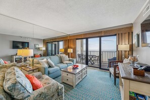 Habitación de hotel con sofá, mesa de centro, televisor, balcón con vistas y escritorio con lámpara.