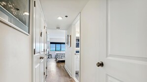 Un couloir avec des portes blanches, un lit recouvert d’une couvre-lit à motifs bleus et blancs, et une fenêtre donnant sur un paysage.